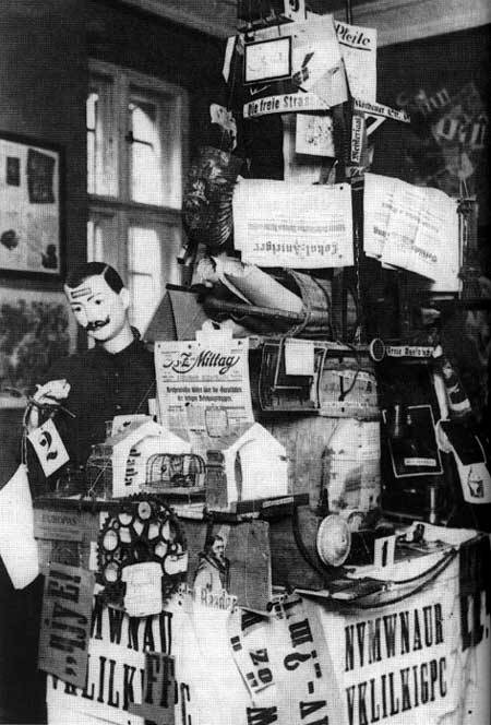 A photograph of an assemblage shown at the Fair. It is made up of newspapers and bric-a-brac, including newspapers and cogs; there is a mustachioed mannequin to the left.