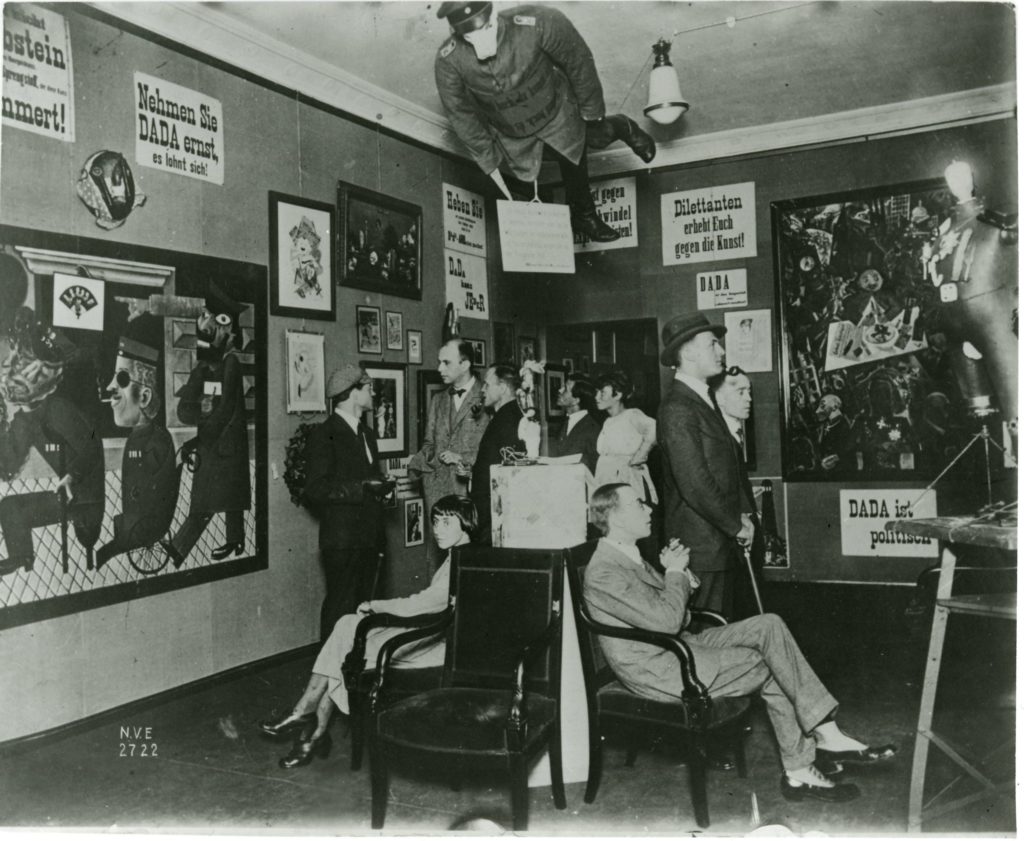 A photograph of the second room of the Dada Fair. A small group of people gathers to look at the work on the walls. There are paintings and posters with Dada slogans in German; above them hangs a mannequin, dressed in soldier's uniform with a pig's head (this is the Prussian Archangel).